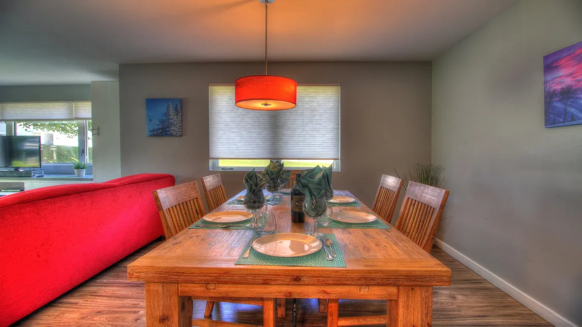 This image features a welcoming dining and living area with a dining table ready for shared meals, a red sofa that invites relaxation, a hanging light fixture, and artwork on the walls. The scene is set for gatherings, celebrations, and quality time among friends and family.