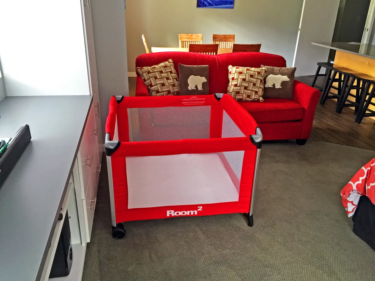 A cozy family-oriented living space featuring a red playpen for children, a red couch, a wooden dining table, and decorative cushions. The setup suggests multi-generational gatherings and casual, comfortable living for groups with varying needs.