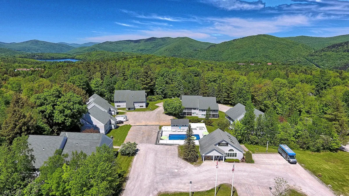 This image captures an exterior scene featuring a swimming pool surrounded by lounge chairs, residential buildings in the background, and a dramatic mountain backdrop. The parking area is also visible, situating the property within a landscape that hints at both relaxation and mountain adventure.