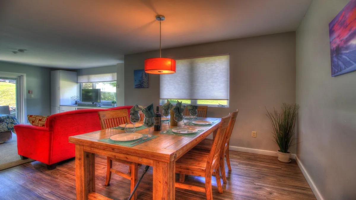 A welcoming dining and lounge area with a dining table set near large windows and a vibrant red sofa. The space is bathed in natural light, emphasizing areas for family meals, sharing stories, and entertaining guests.