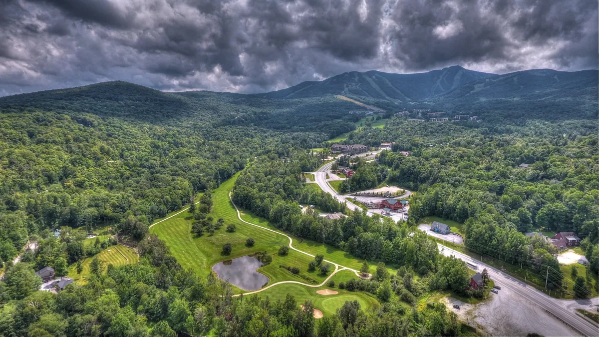 This scenic view captures lush green mountains, a winding river, a well-manicured golf course, and a small pond. The image evokes the tranquility of Vermont's outdoors with inviting spaces for hiking, golfing, and nature exploration.