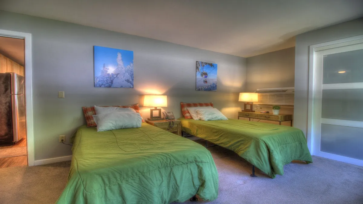 A cozy interior featuring two twin beds with crisp, green bedspreads, complemented by matching lamps on the nightstand and calming wall art. The beds are spaced apart, creating an inviting space for friends or family members who prefer separate sleeping arrangements.