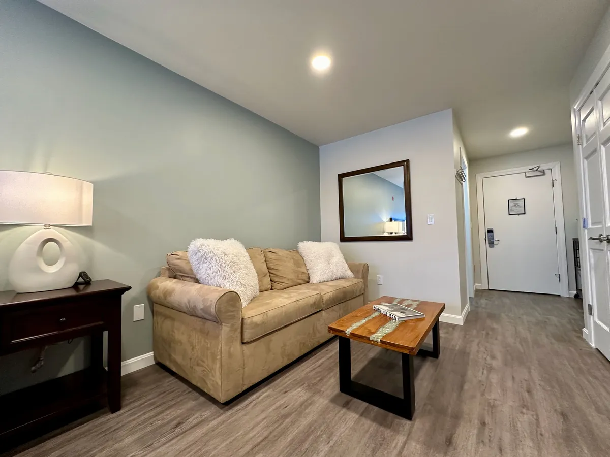 A welcoming entryway with a sofa adorned with cushions, a wooden coffee table, a lamp, a mirror, and the entrance door in view. The mood is cozy and inviting, suggesting a warm welcome upon arrival and ease of access.