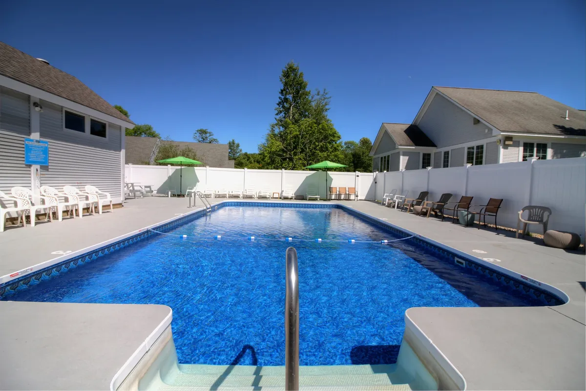 A beautiful outdoor heated saltwater pool surrounded by lush greenery, lounge chairs, and green umbrellas. The scene suggests a relaxed, inviting atmosphere perfect for swimming, sunbathing, or poolside conversations. The pool is the focal point, with trees providing a natural Vermont backdrop.