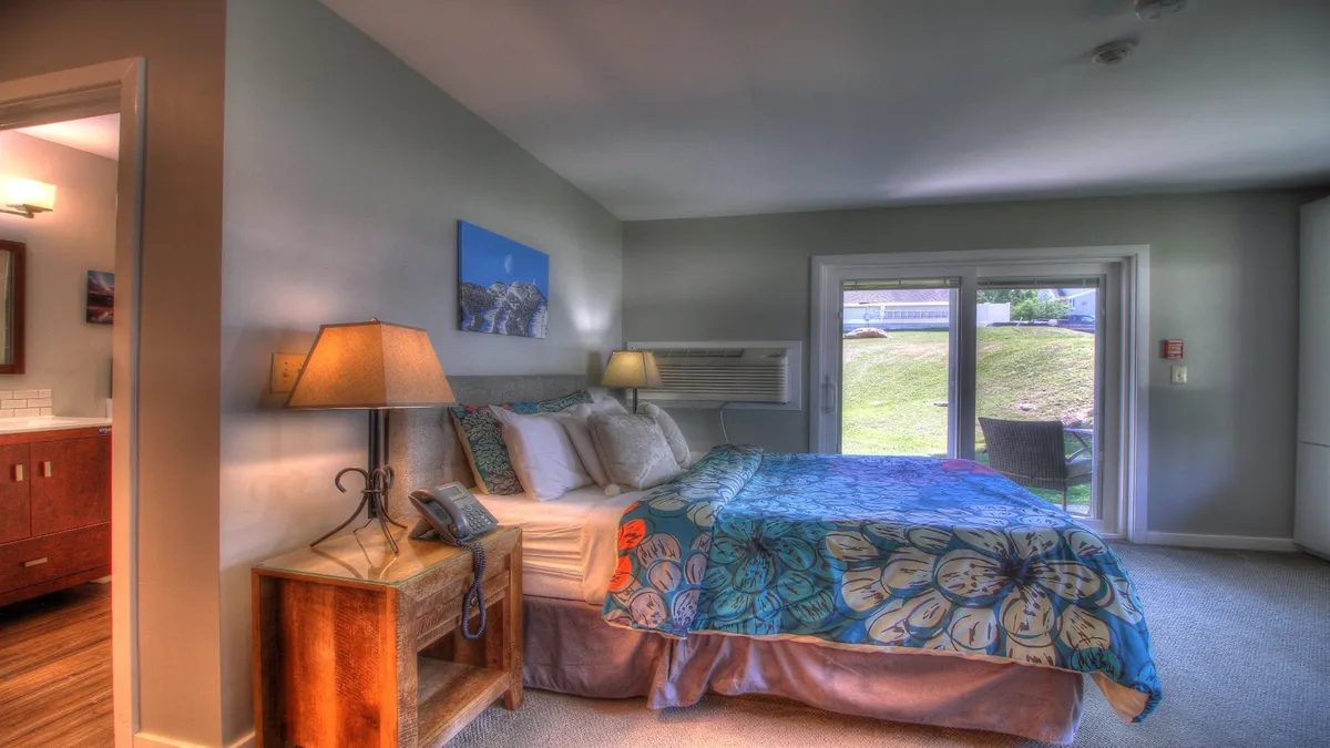 A comfortable bedroom with floral patterned bedding is anchored by sliding glass doors that open to an outdoor view, inviting abundant natural light. A nightstand with a warm lamp sits beside the bed, suggesting cozy mornings and easy access to the outdoors.