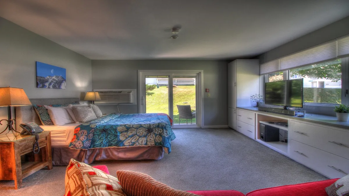 A serene bedroom with a comfortable bed, a television, and an armchair faces a sliding glass door that opens onto a balcony or patio. The space is flooded with soft, natural light, offering a tranquil spot for morning coffee while enjoying the landscape beyond.