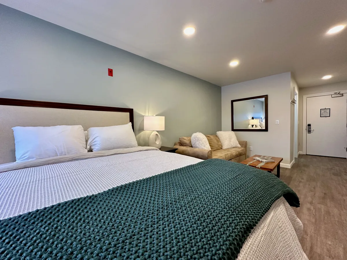 A tranquil bedroom scene with a plush bed draped in a green throw blanket, a cozy sofa, a small table with magazines, and a large mirror reflecting the inviting atmosphere. The soft lighting and comfortable furnishings evoke a restful, hotel-like retreat.