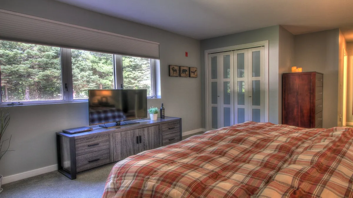 A cozy bedroom with a plaid comforter, a television on a wooden console, and large windows overlooking leafy trees. Decorative wall art and a wooden dresser add warmth, while the view outside the windows hints at the picturesque Vermont setting.