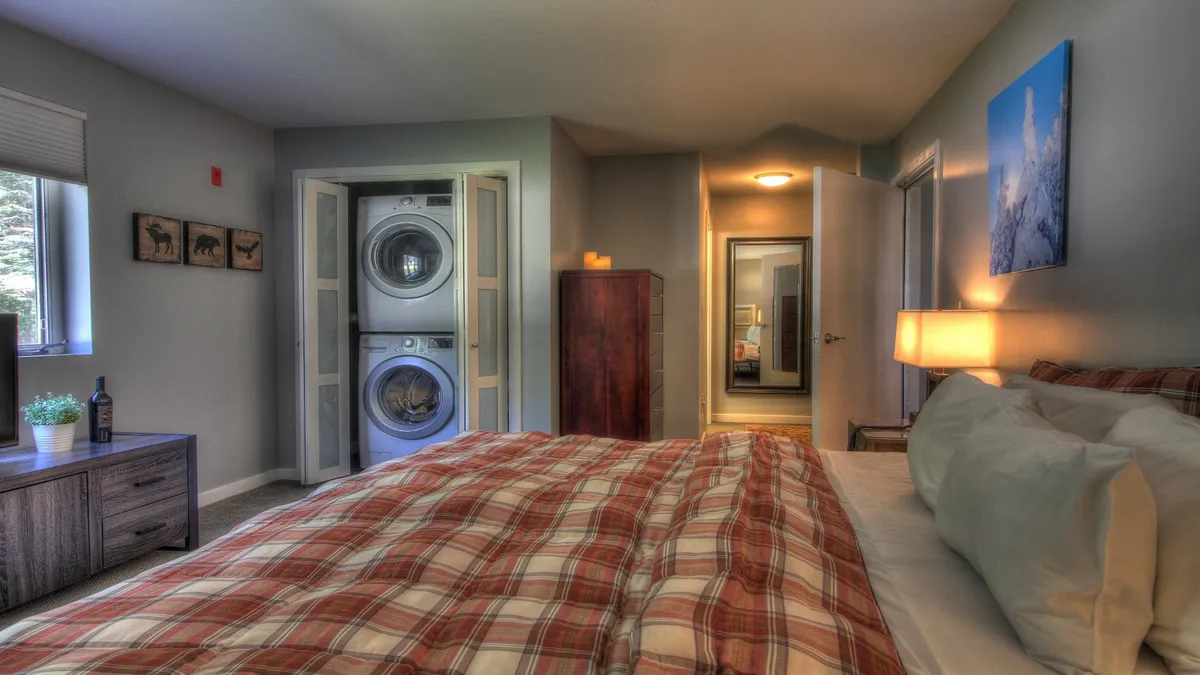 This image captures a cozy bedroom with plaid bedding, a nightstand with a lamp, and prominently features a stacked washer and dryer unit in the corner. The space feels inviting and functional, blending comfort and practical amenities.