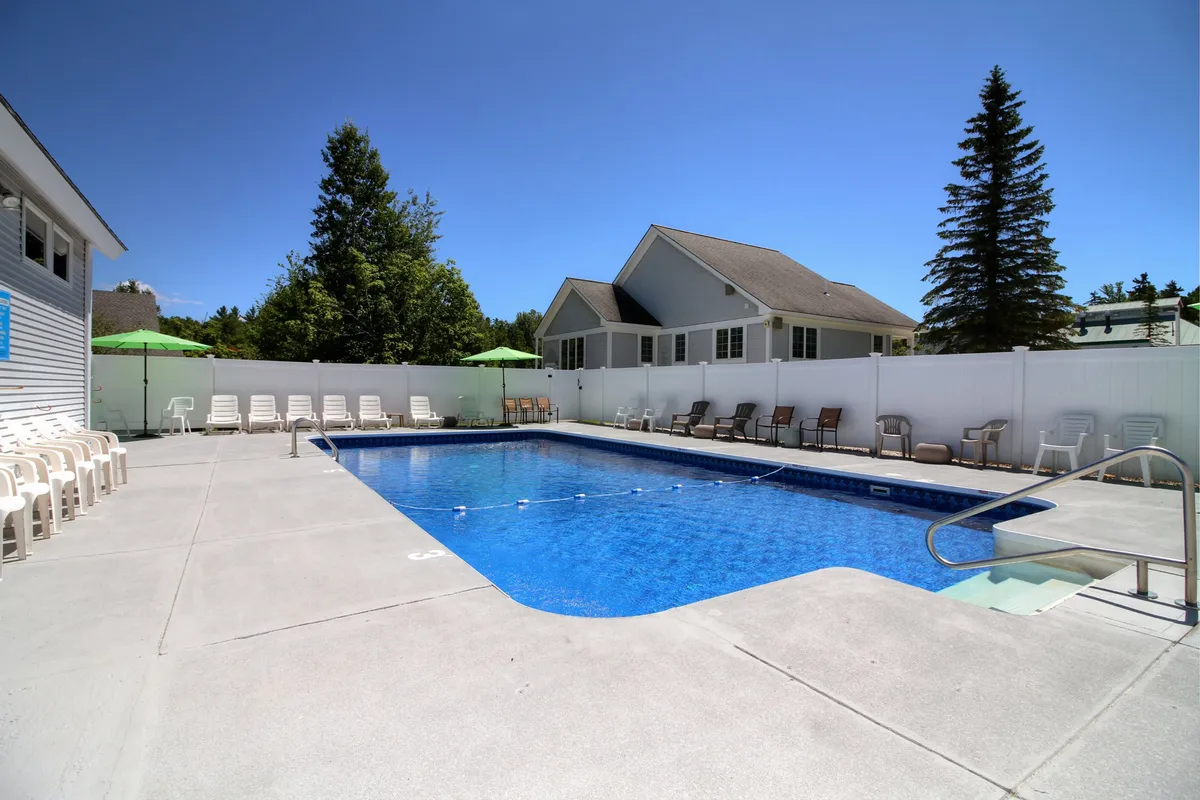 This image showcases a sparkling outdoor swimming pool surrounded by lounge chairs and green umbrellas, all set amidst lush trees within a fenced area. The relaxed atmosphere is ideal for both swimming and socializing, with plenty of space for families and groups to unwind under the sun.
