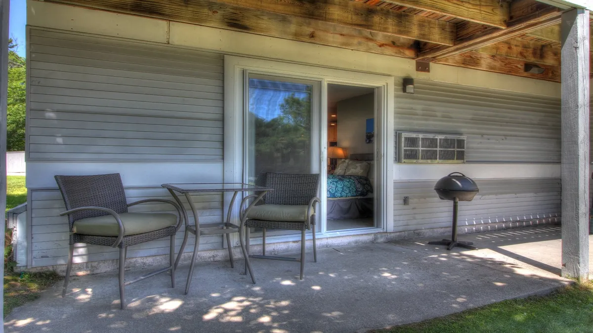 A close-up exterior shot of a patio area with chairs, a table, and a grill next to a sliding glass door. The setup suggests a casual, cozy environment for outdoor dining, grilling, or enjoying evening conversations as the sun sets.