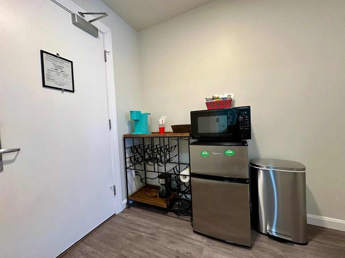 A compact kitchenette area with a microwave, mini fridge, shelf, and nearby door, designed for handy snack and drink preparation. The scene feels convenient and organized, ideal for quick refreshments and storing complimentary bottled water.